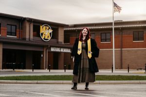 Graduate in cap and gown smiling outside Watkins Memorial High School with American flag.