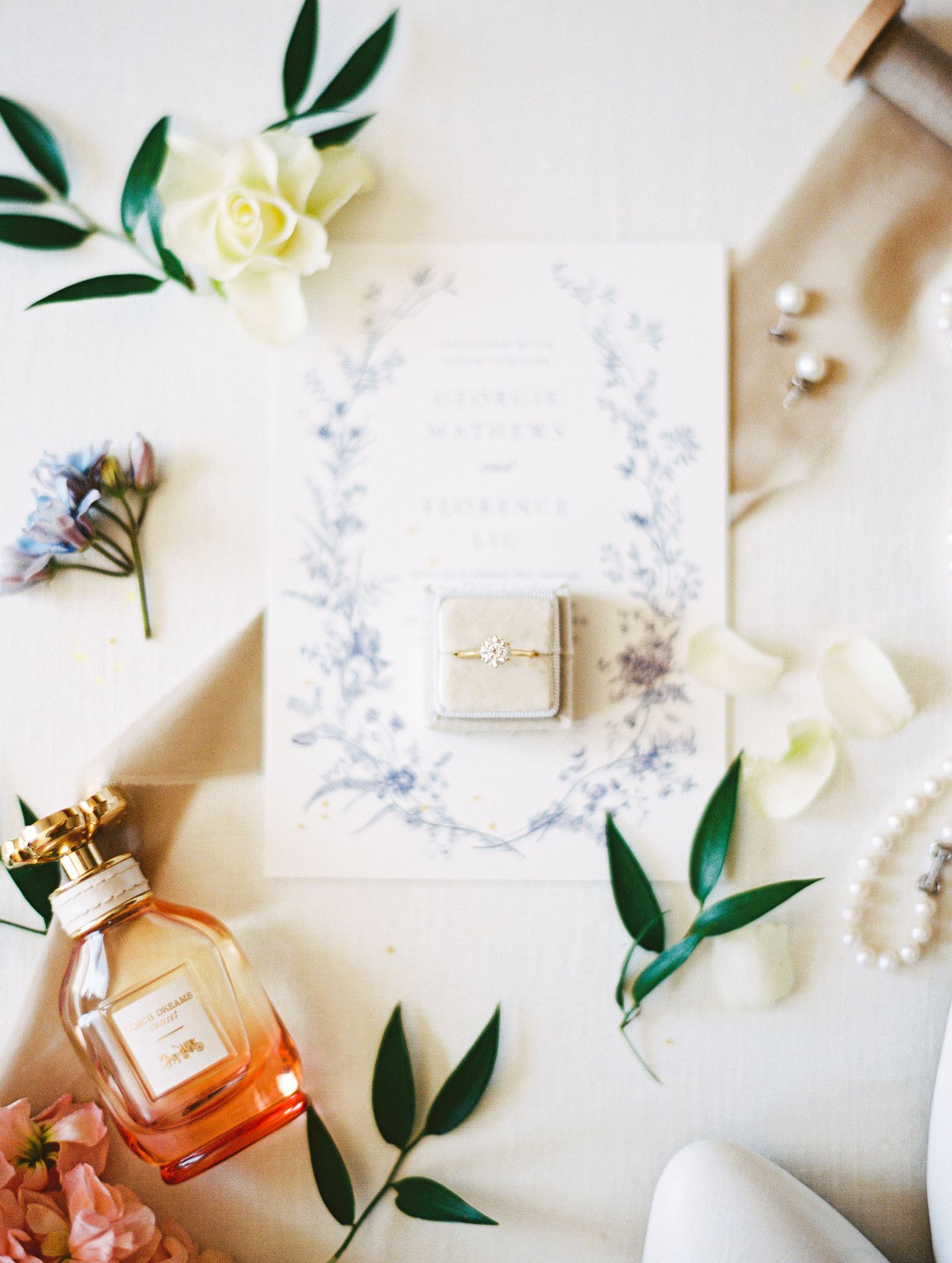 Elegant wedding flat lay with blue floral invitation, diamond ring, perfume bottle, pearl bracelet, and white flowers.