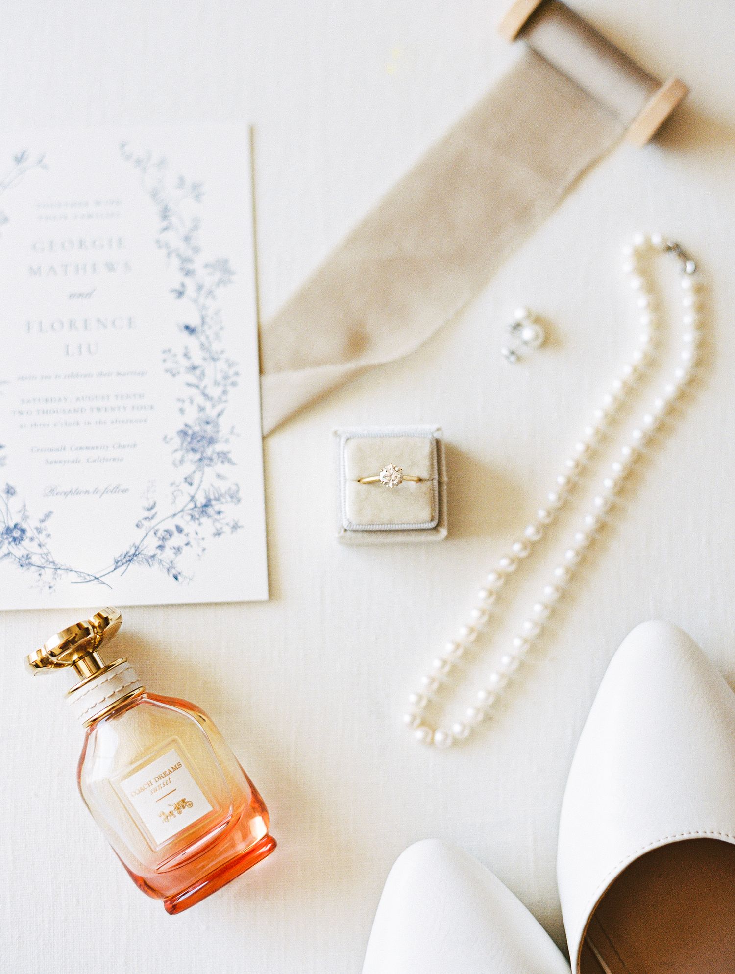 Elegant wedding flat lay featuring floral invitation, pearl necklace, engagement ring, perfume, and bridal accessories.