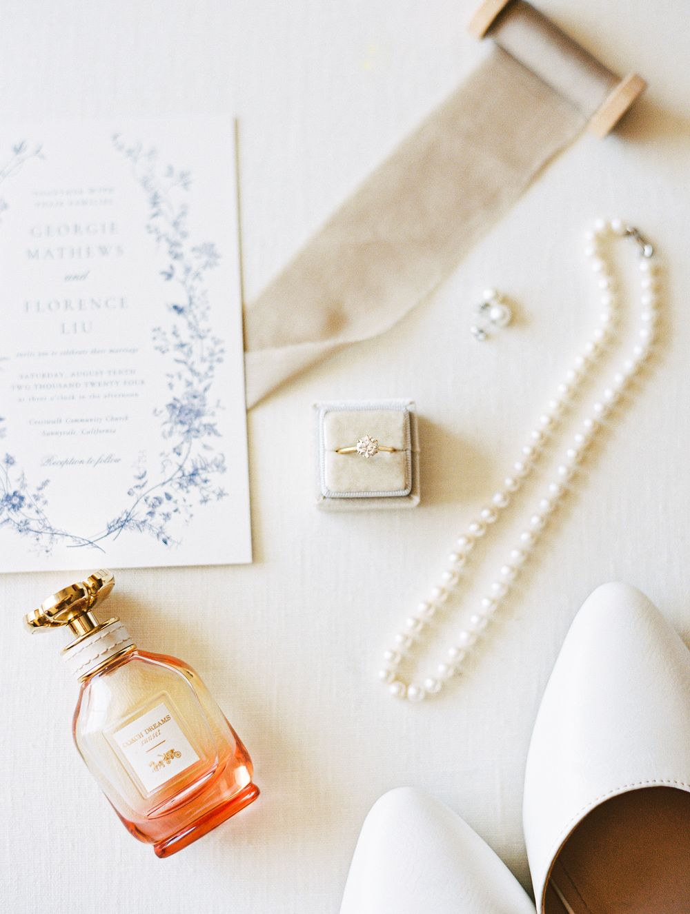 Elegant wedding flat lay featuring floral invitation, pearl necklace, engagement ring, perfume, and bridal accessories.