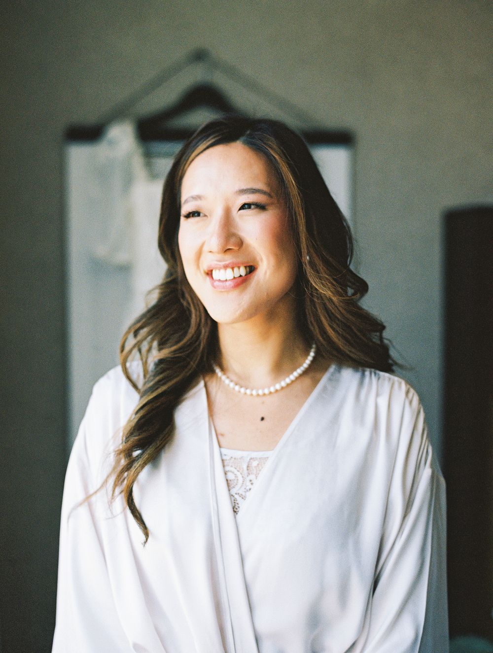 Smiling bride wearing pearl necklace and white robe with wedding dress hanging in background during bridal preparations.
