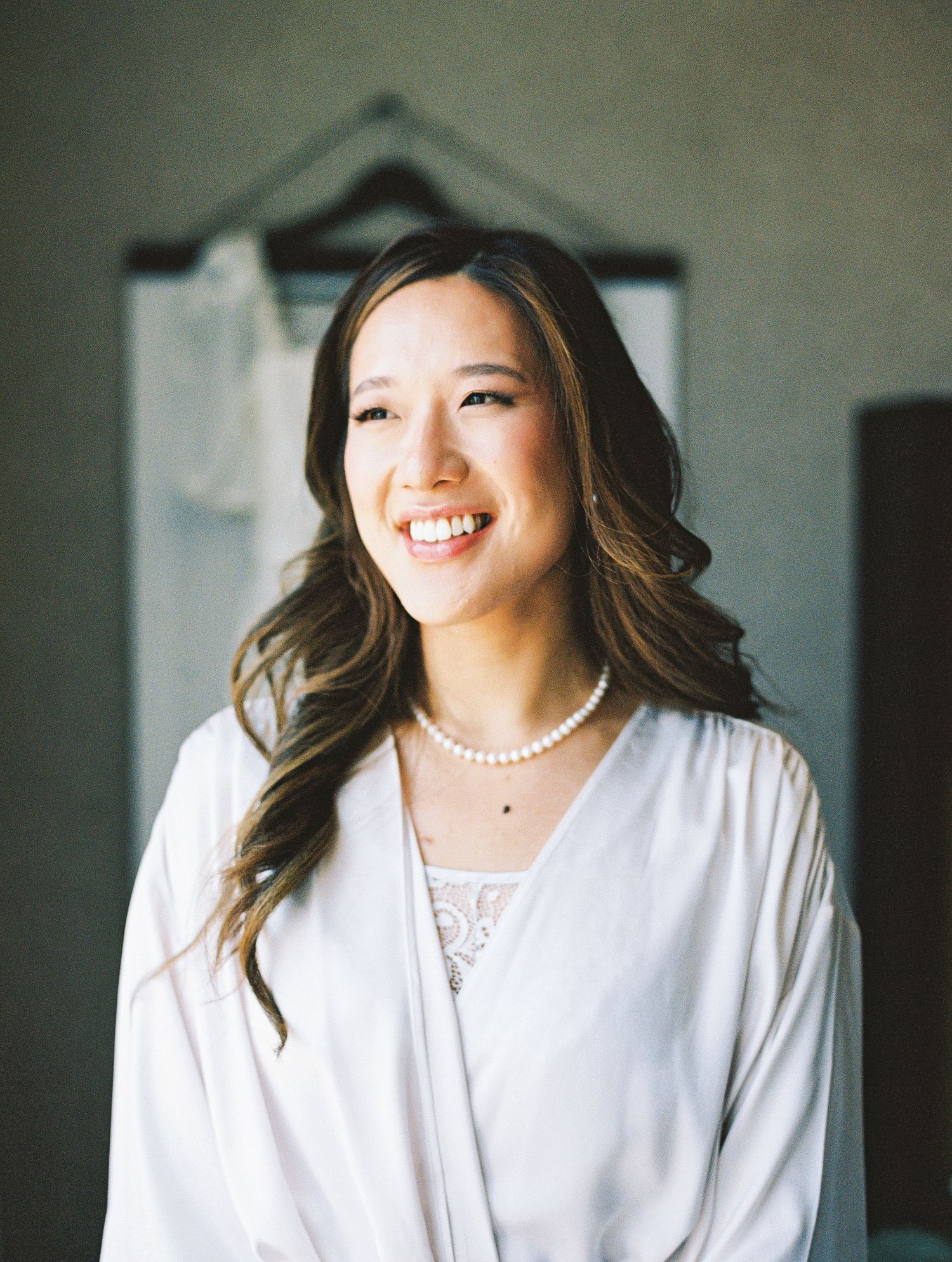 Smiling bride wearing pearl necklace and white robe with wedding dress hanging in background during bridal preparations.