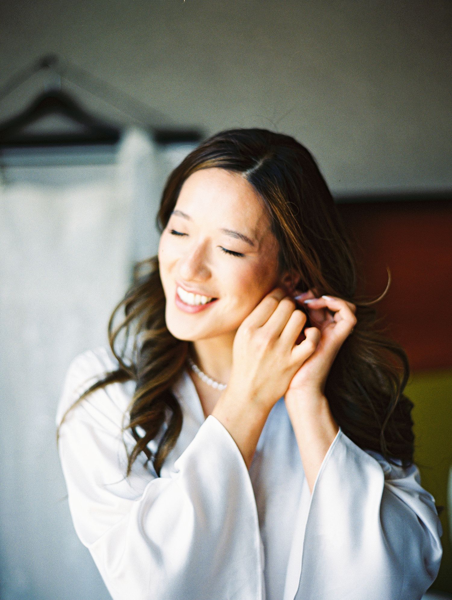 Smiling bride in white robe putting on pearl earrings while getting ready on her wedding day.