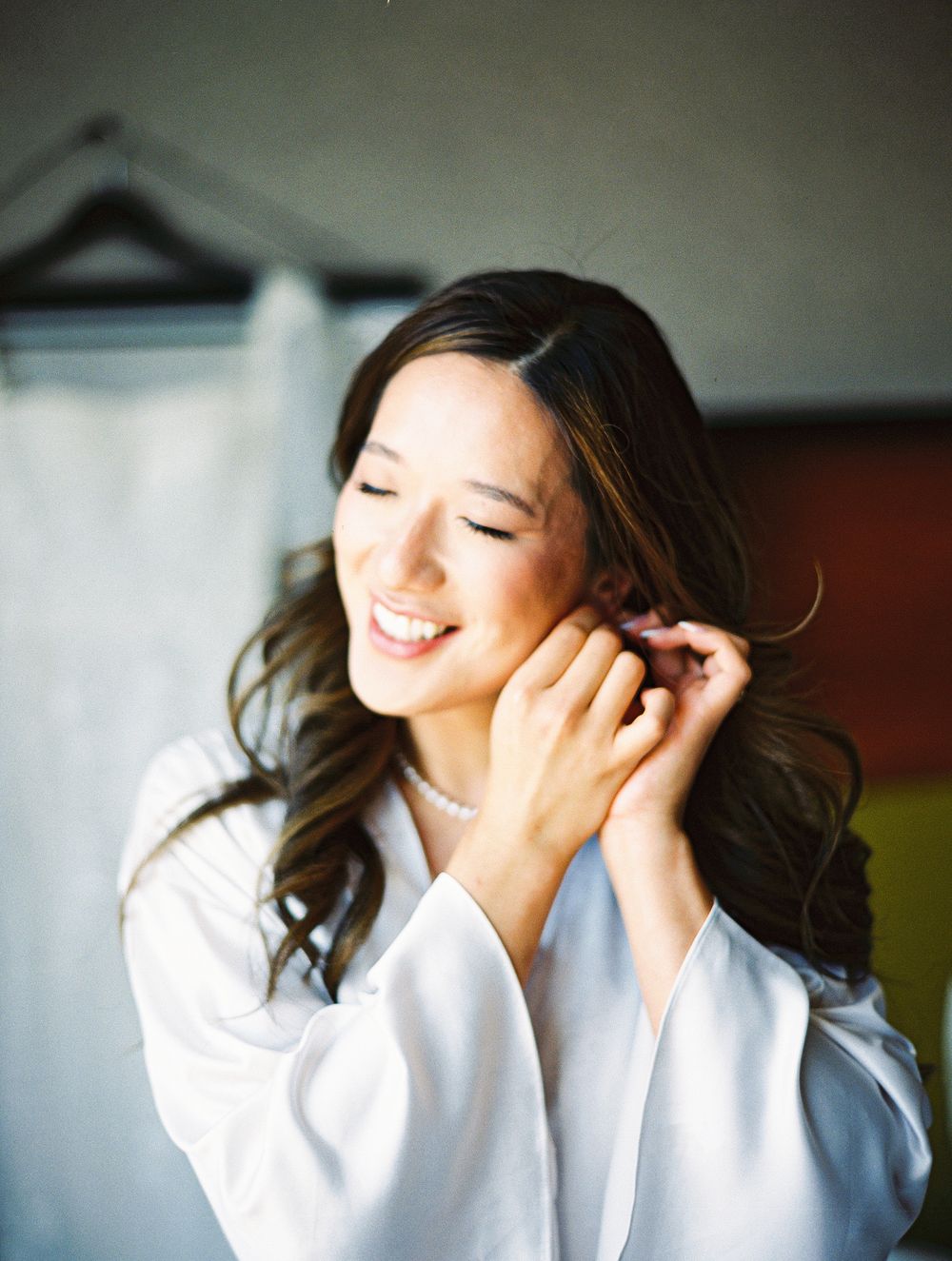 Smiling bride in white robe putting on pearl earrings while getting ready on her wedding day.