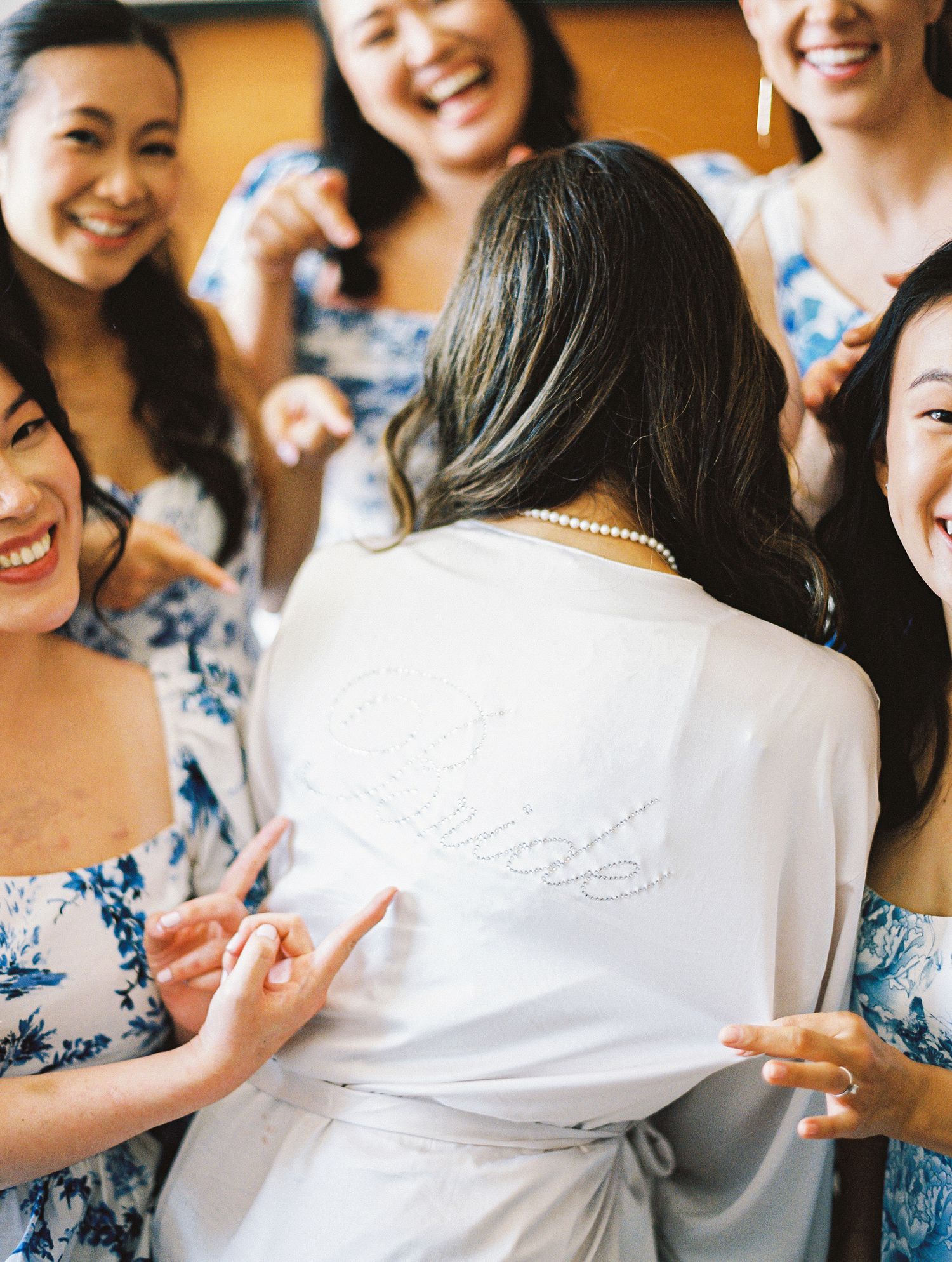 Bride in monogrammed white robe surrounded by laughing bridesmaids in blue floral dresses.