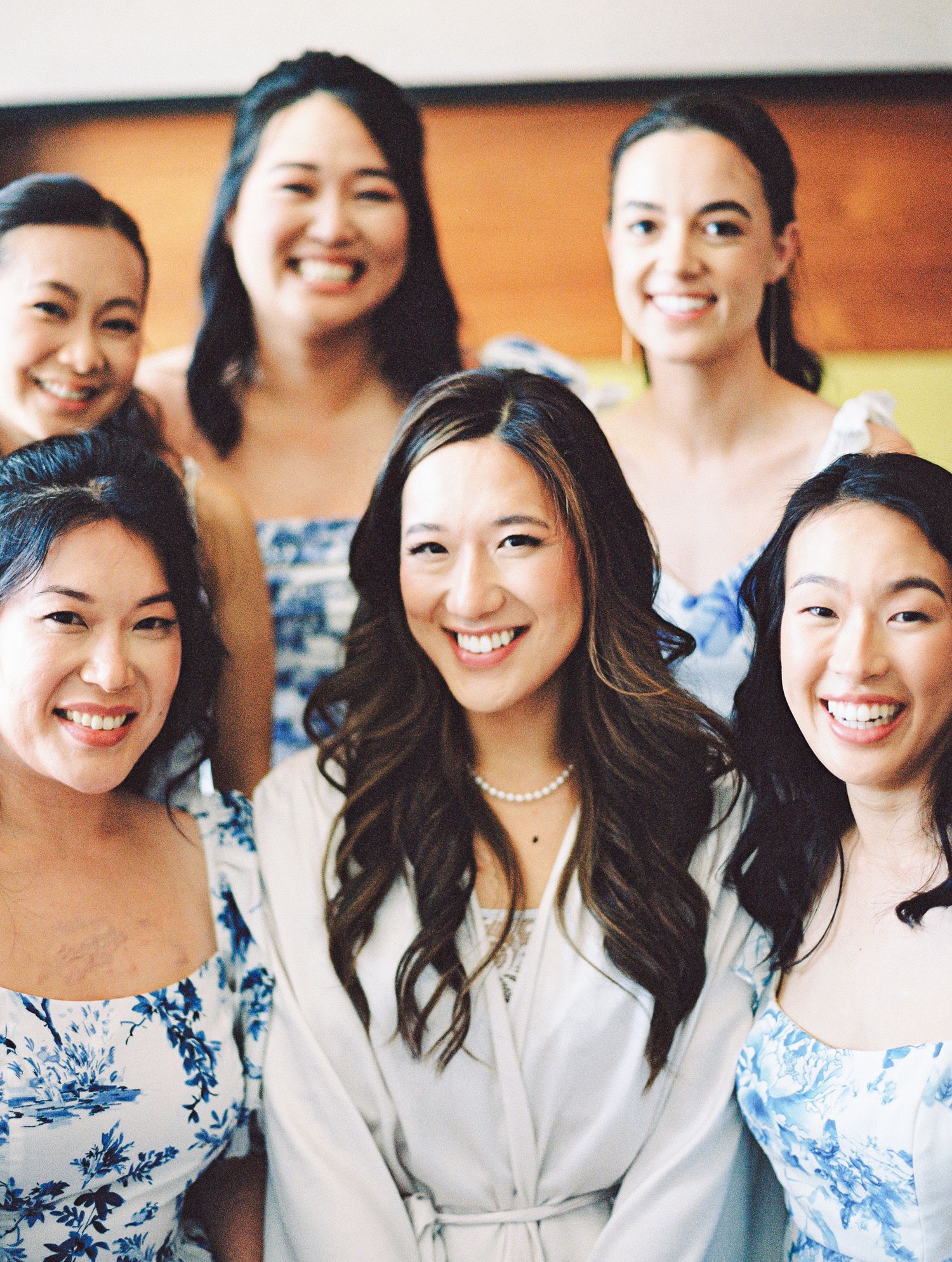Bride in white robe smiles with bridesmaids in blue floral dresses during wedding preparation.
