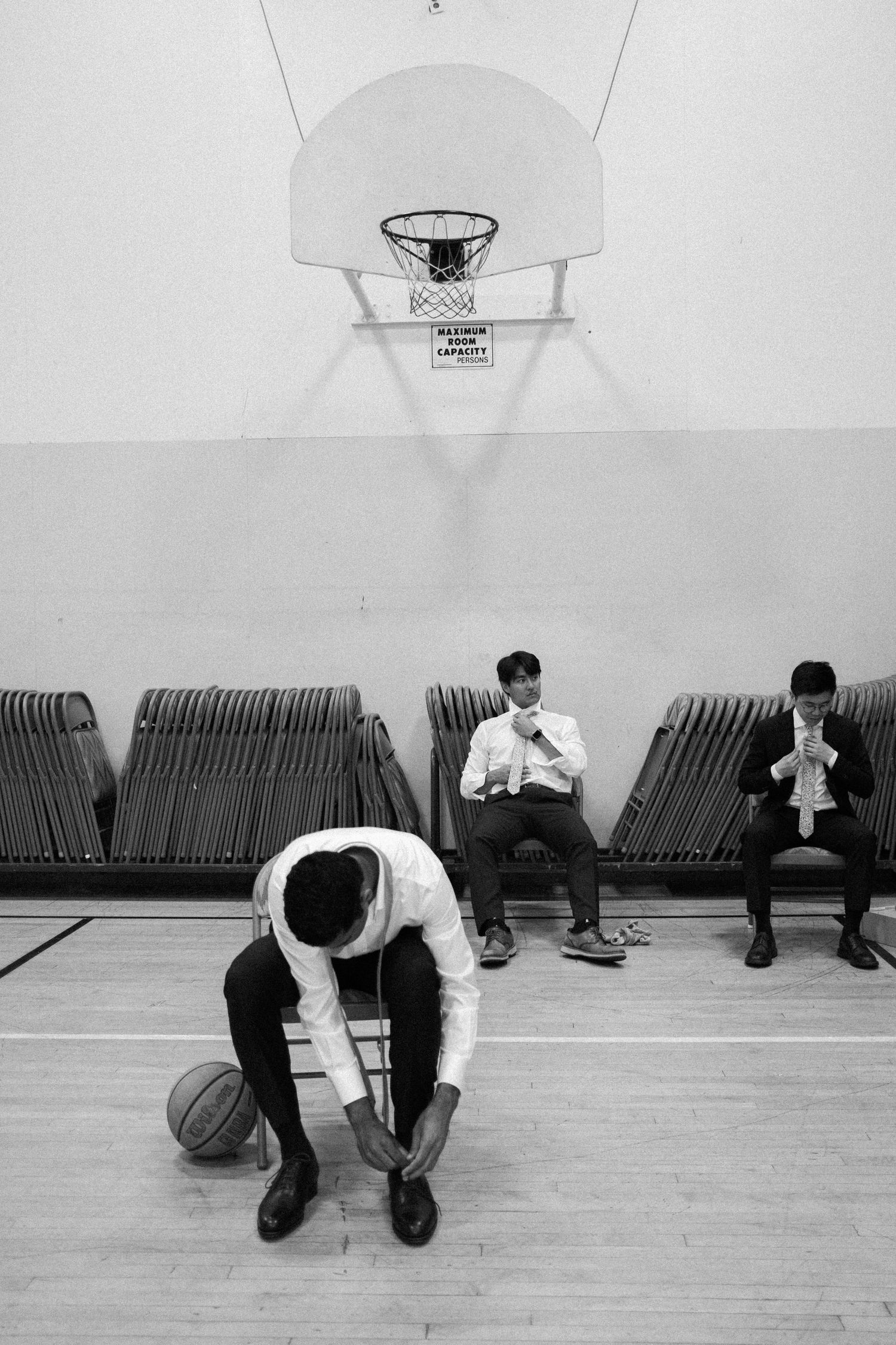 Black and white photo of groomsmen preparing in a gymnasium, one tying shoes near a basketball.