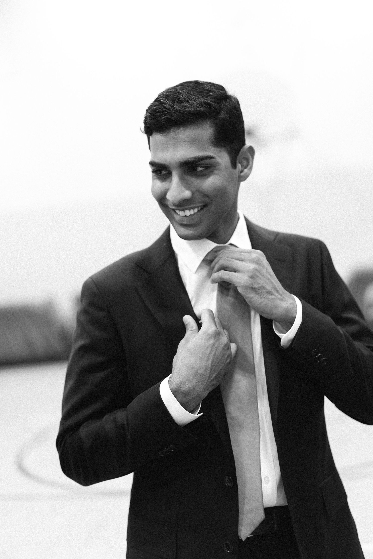 Smiling groom in black suit adjusting his tie outdoors in black and white photo.