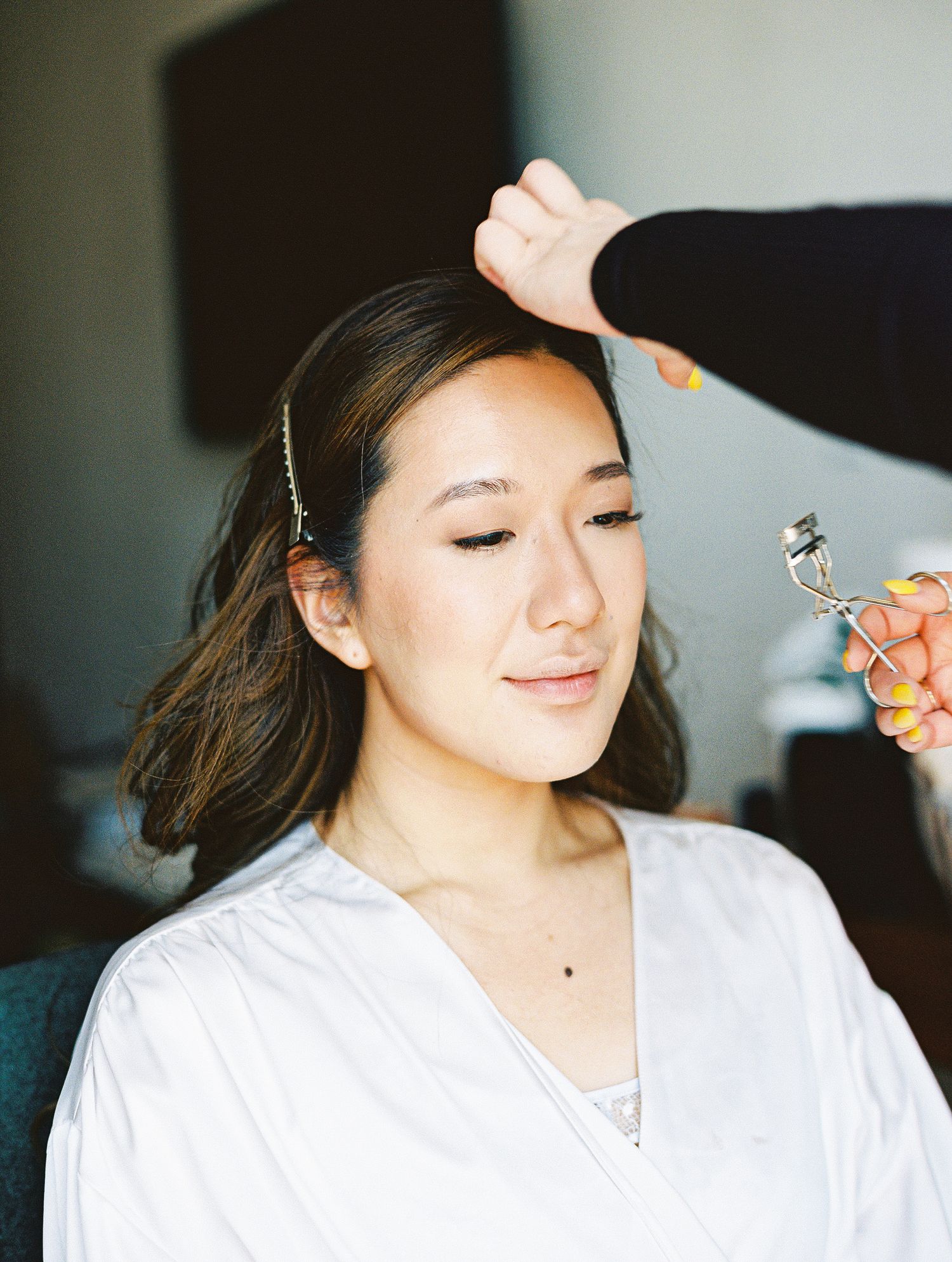 A makeup artist curling a young Asian woman's eyelashes while styling her for a special occasion.