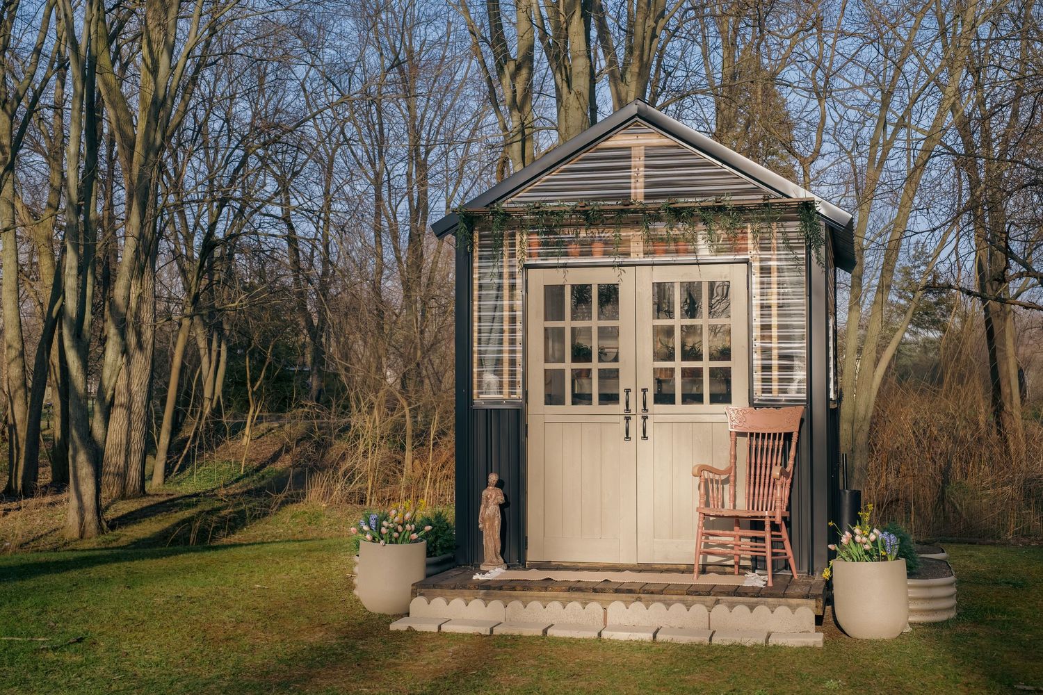 Charming small garden shed with French doors, rocking chair, and potted plants surrounded by bare trees in a serene backyard.