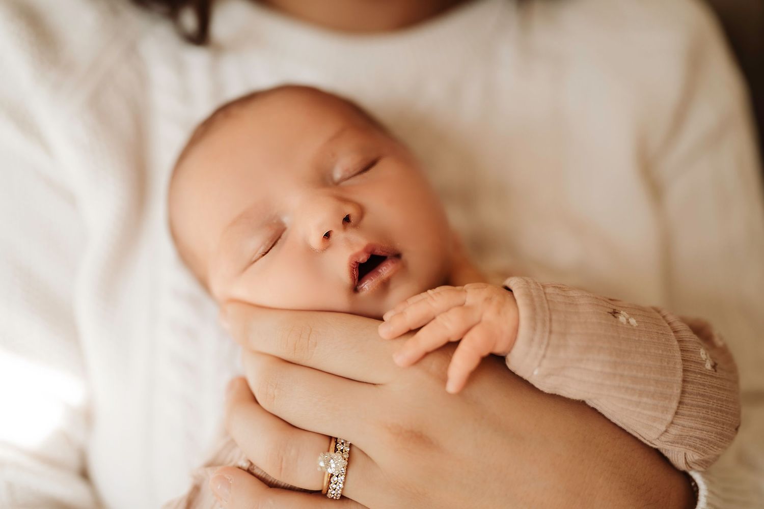 Peaceful newborn baby sleeping in mother's arms, wearing a soft pink onesie, with delicate tiny fingers visible.