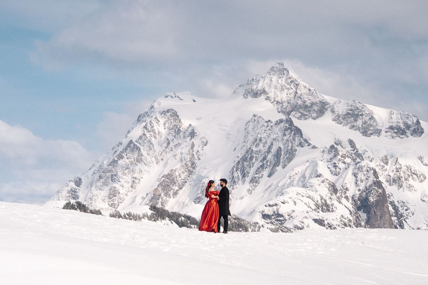 Couple in red dress embracing on snowy mountain landscape with dramatic snow-capped peaks backdrop.