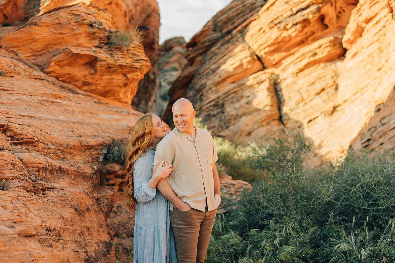 Couple embracing among red rock canyon formations with lush greenery, bathed in warm golden sunset light.
