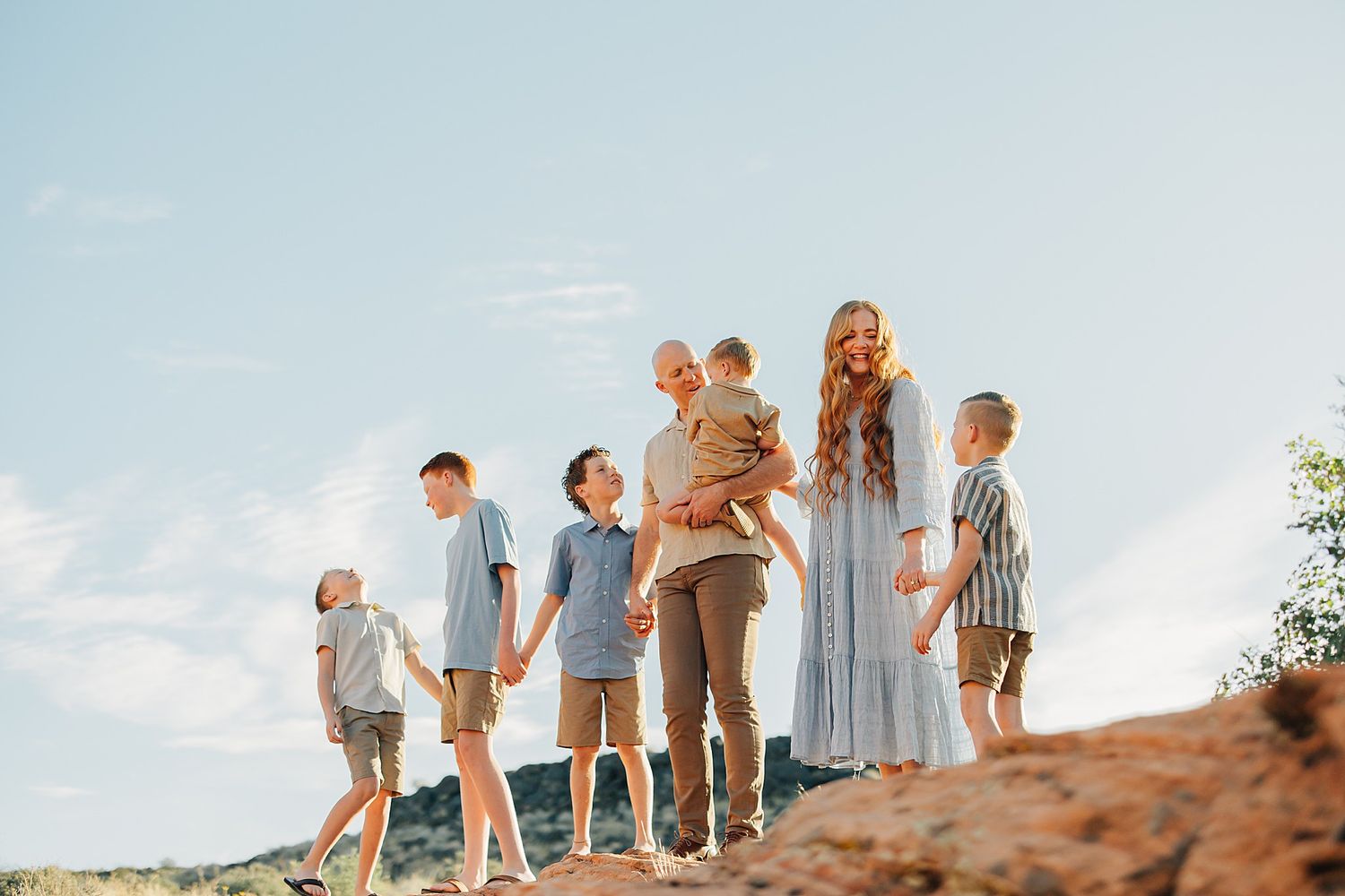 Family of six poses on red rock terrain under blue sky, mother in flowing dress holds toddler while children hold hands.