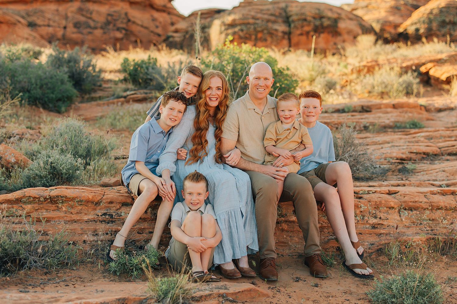 Family of six poses on red rock ledge in desert landscape, parents with four children in coordinating blue and tan outfits.