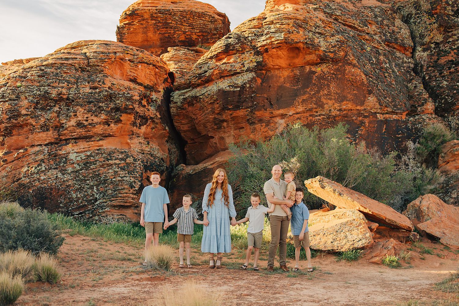 Family of six posing in front of red rock formations in an arid desert landscape during golden hour.