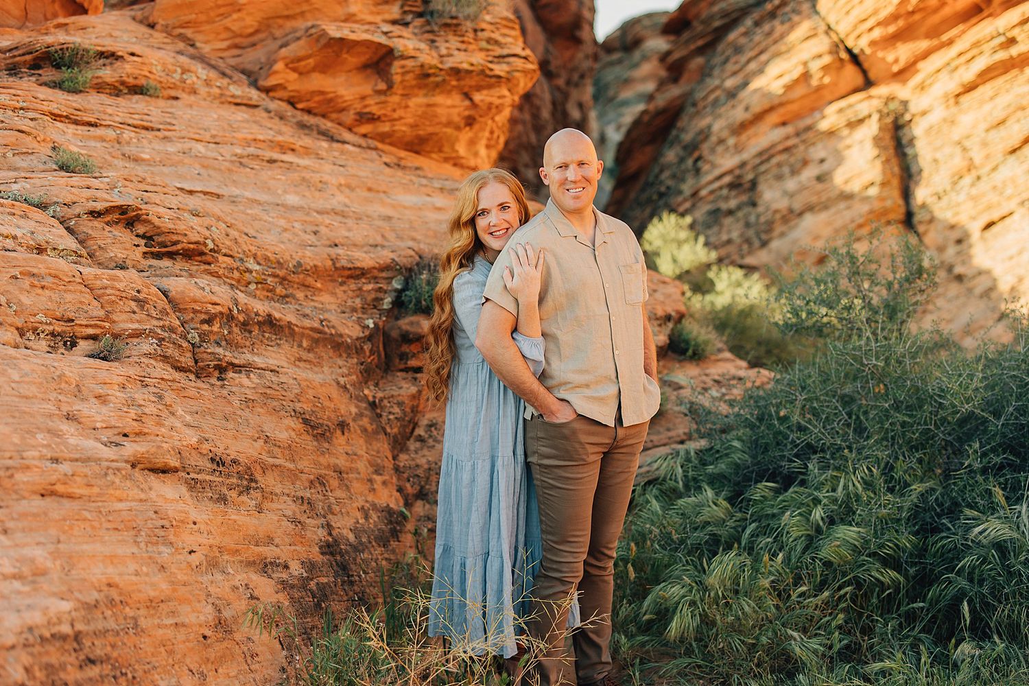 Couple posing together against red rock canyon walls in golden hour sunlight, woman in blue dress and man in khaki.