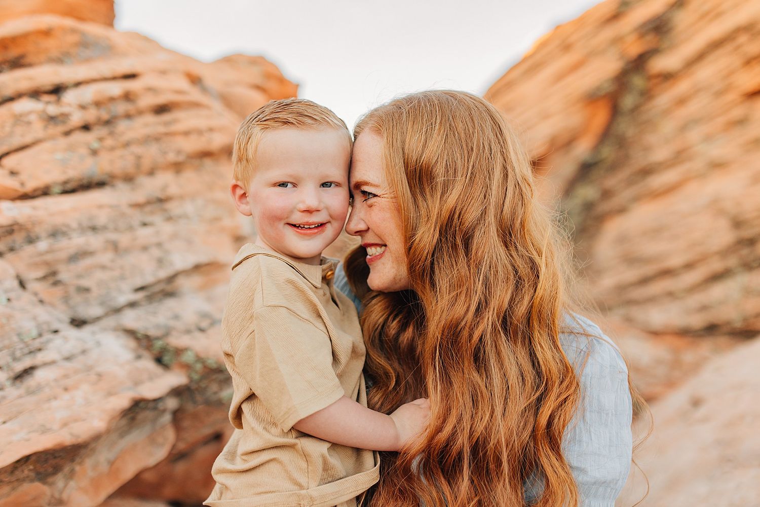 A mother with long red wavy hair holds her smiling young blond son against red rock canyon walls.