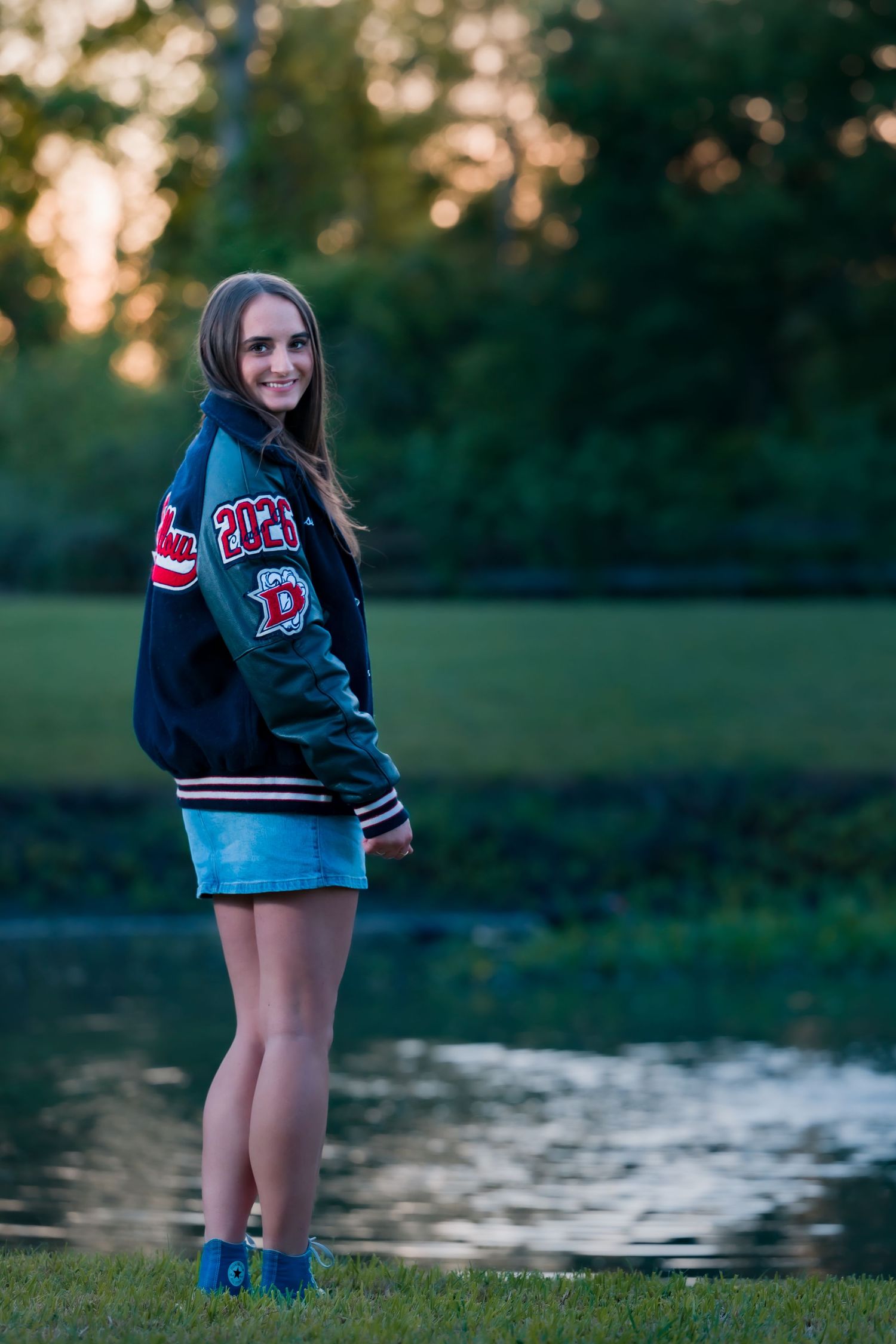 Teen girl in varsity jacket and denim skirt poses by a pond at sunset with trees in background.