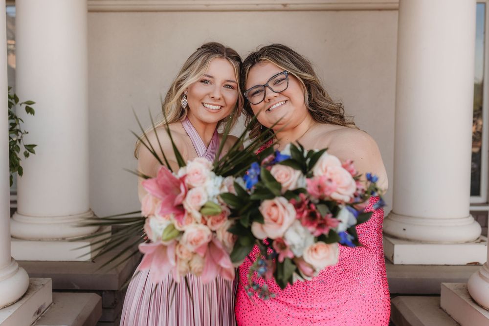 Two smiling women in pink dresses holding floral bouquets pose between white columns.