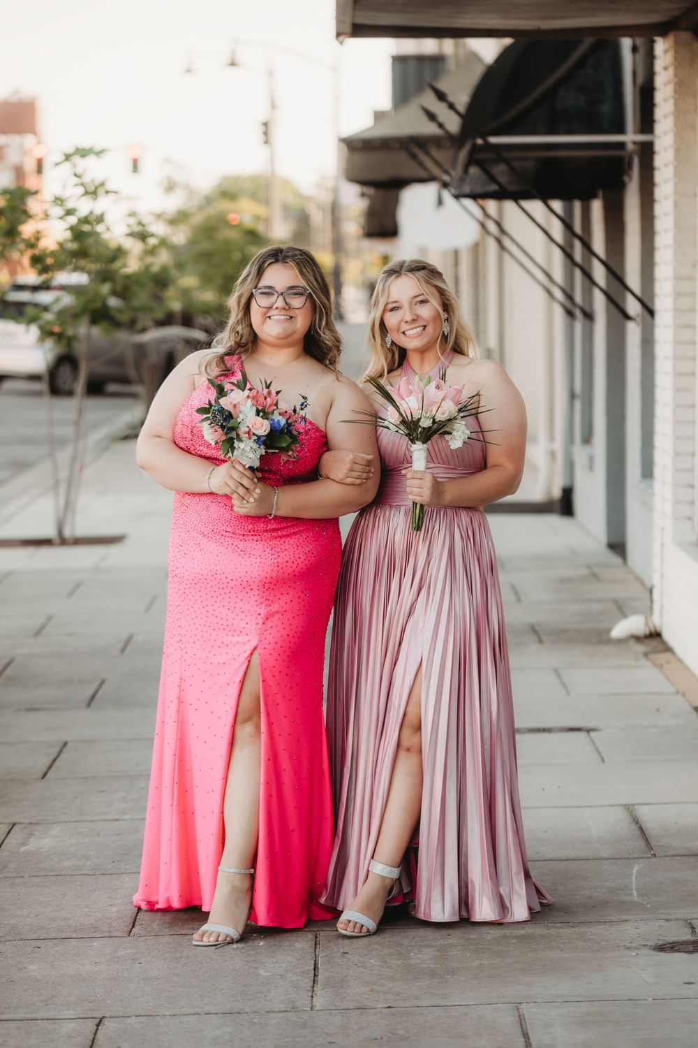 Two bridesmaids in pink gowns holding bouquets pose on a city sidewalk during an outdoor wedding celebration.