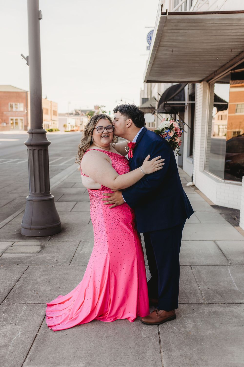 Couple poses on urban sidewalk, woman in pink sequin gown receives kiss from man in navy suit.