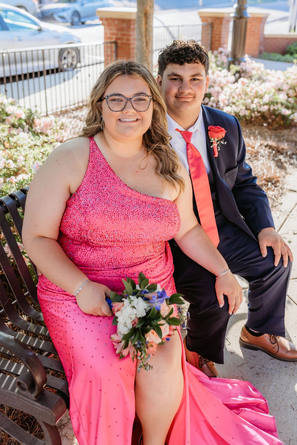 Couple in prom attire sitting on bench, woman in pink gown holding bouquet, man in navy suit with orange boutonniere.