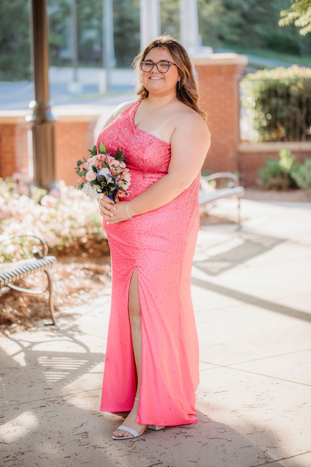 Young woman in pink sequin gown holding floral bouquet poses outdoors for prom photos.