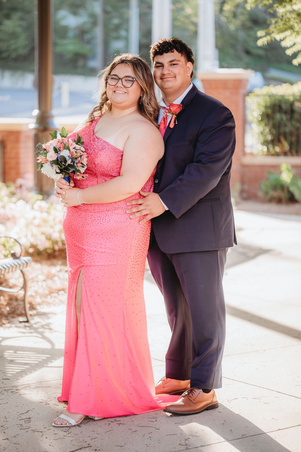 A couple poses in formal prom attire, she in a coral sequin gown holding a bouquet, he in a navy suit with coral bow tie.