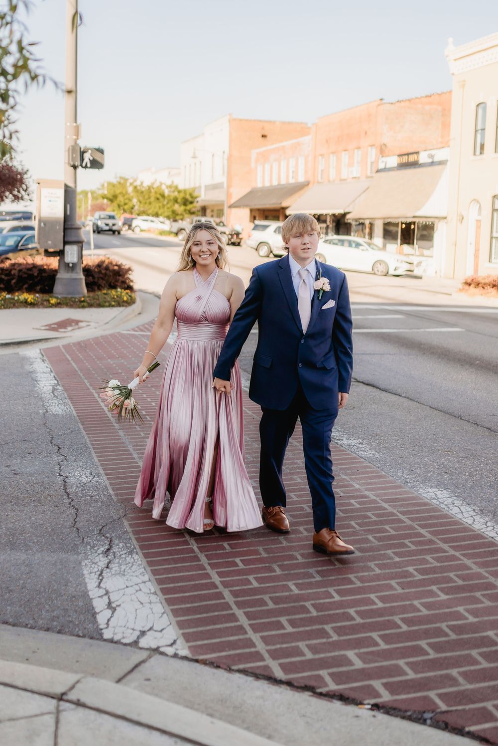 A young couple in formal attire walks hand-in-hand on a brick sidewalk, she in a pink gown, he in a navy suit.