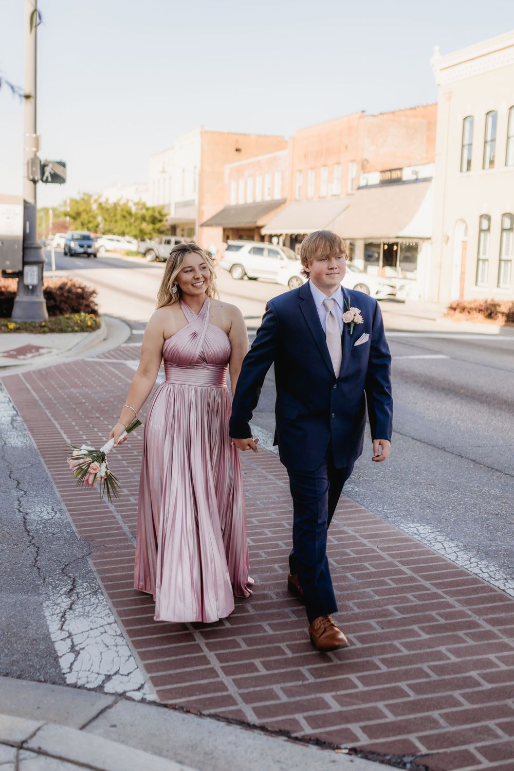 Prom couple walks hand-in-hand across brick crosswalk, woman in pink pleated gown holding bouquet.