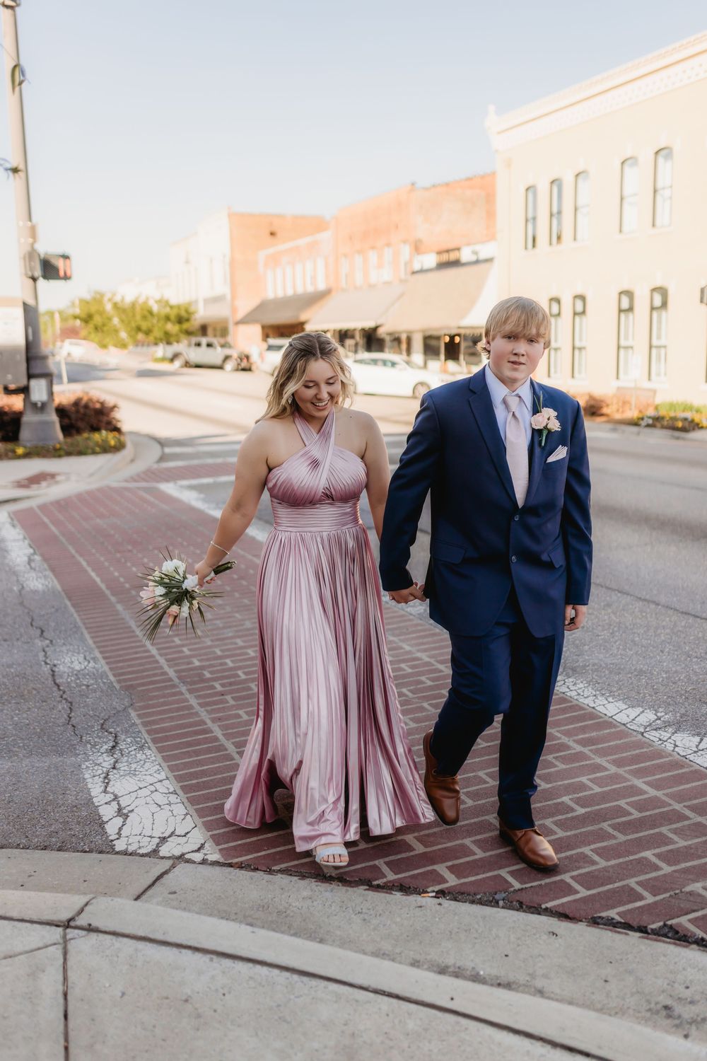 Prom couple walking hand-in-hand across brick crosswalk, woman in mauve gown, man in navy suit with boutonniere.