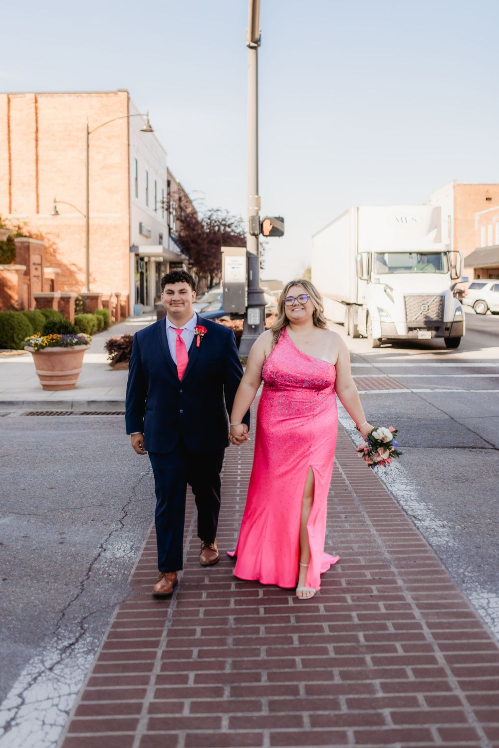 Couple in prom attire walking hand-in-hand on a brick sidewalk, woman in pink gown, man in navy suit.