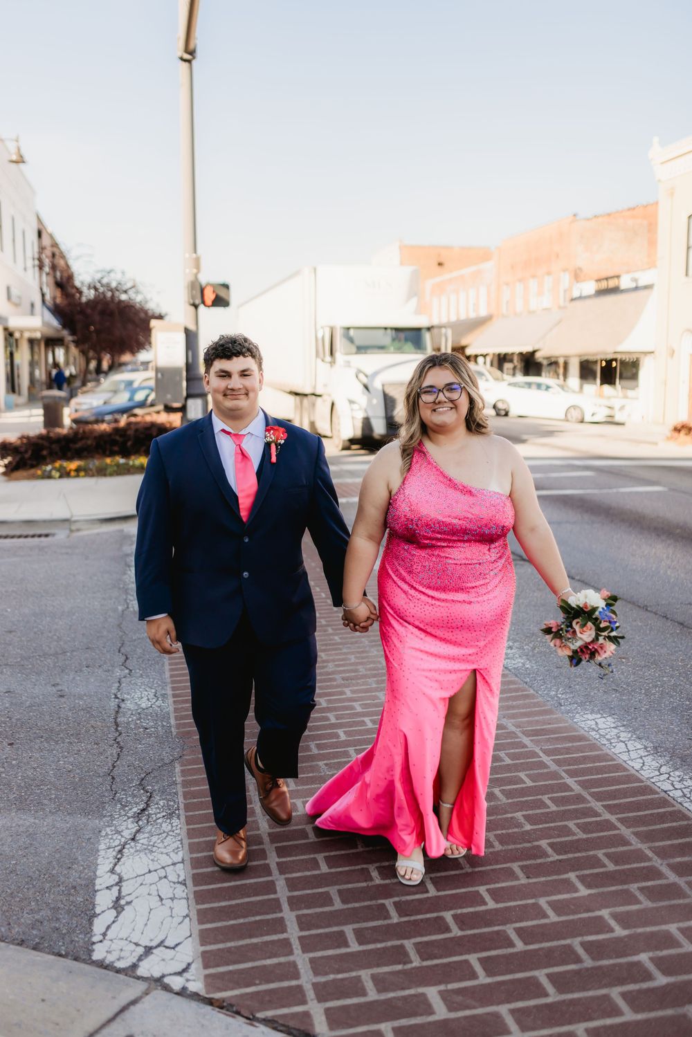 Couple in prom attire walking hand-in-hand, man in navy suit, woman in pink gown, on a brick crosswalk downtown.