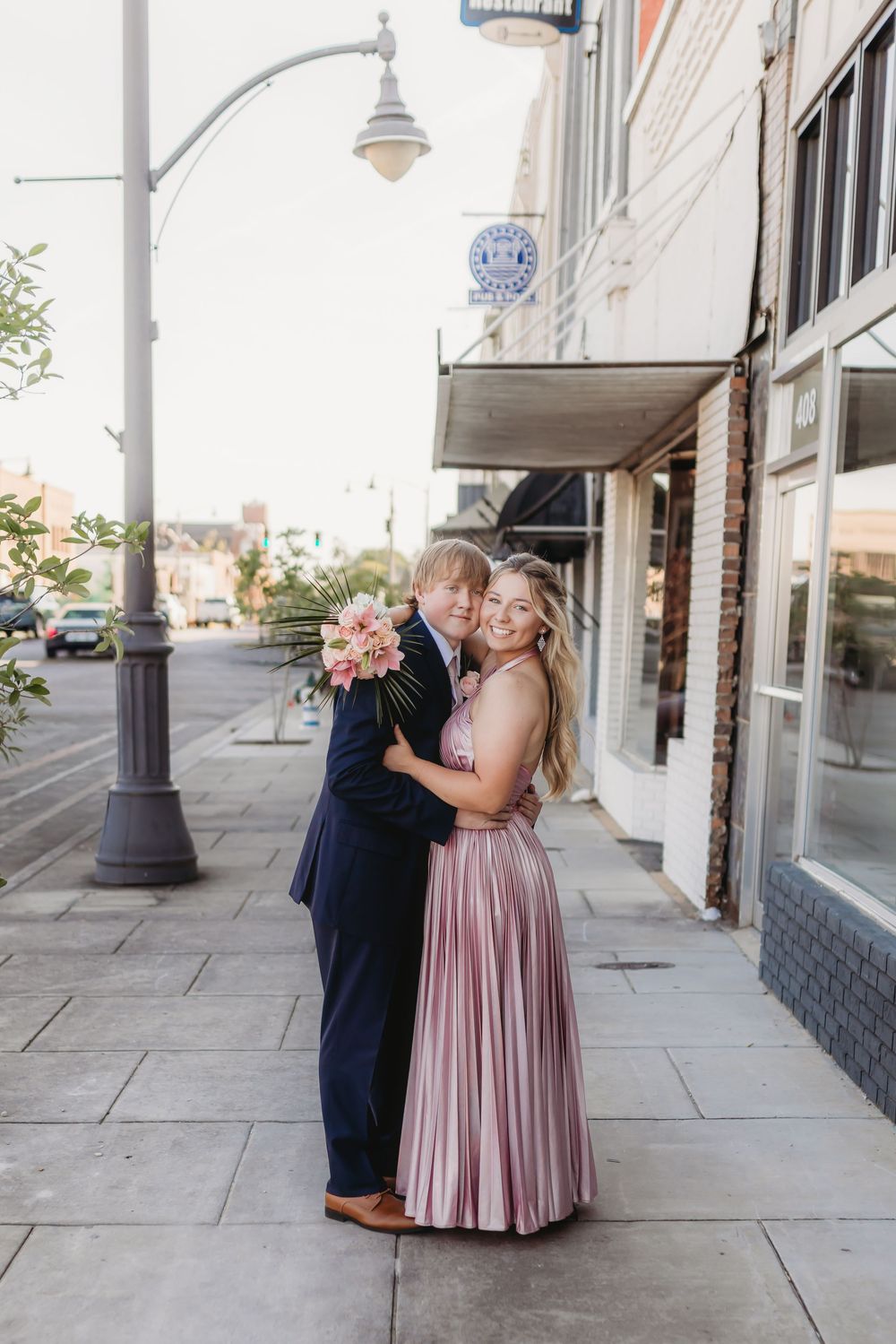 Couple in formal wear embracing on a downtown sidewalk, woman in pink pleated gown holding bouquet, man in navy suit.