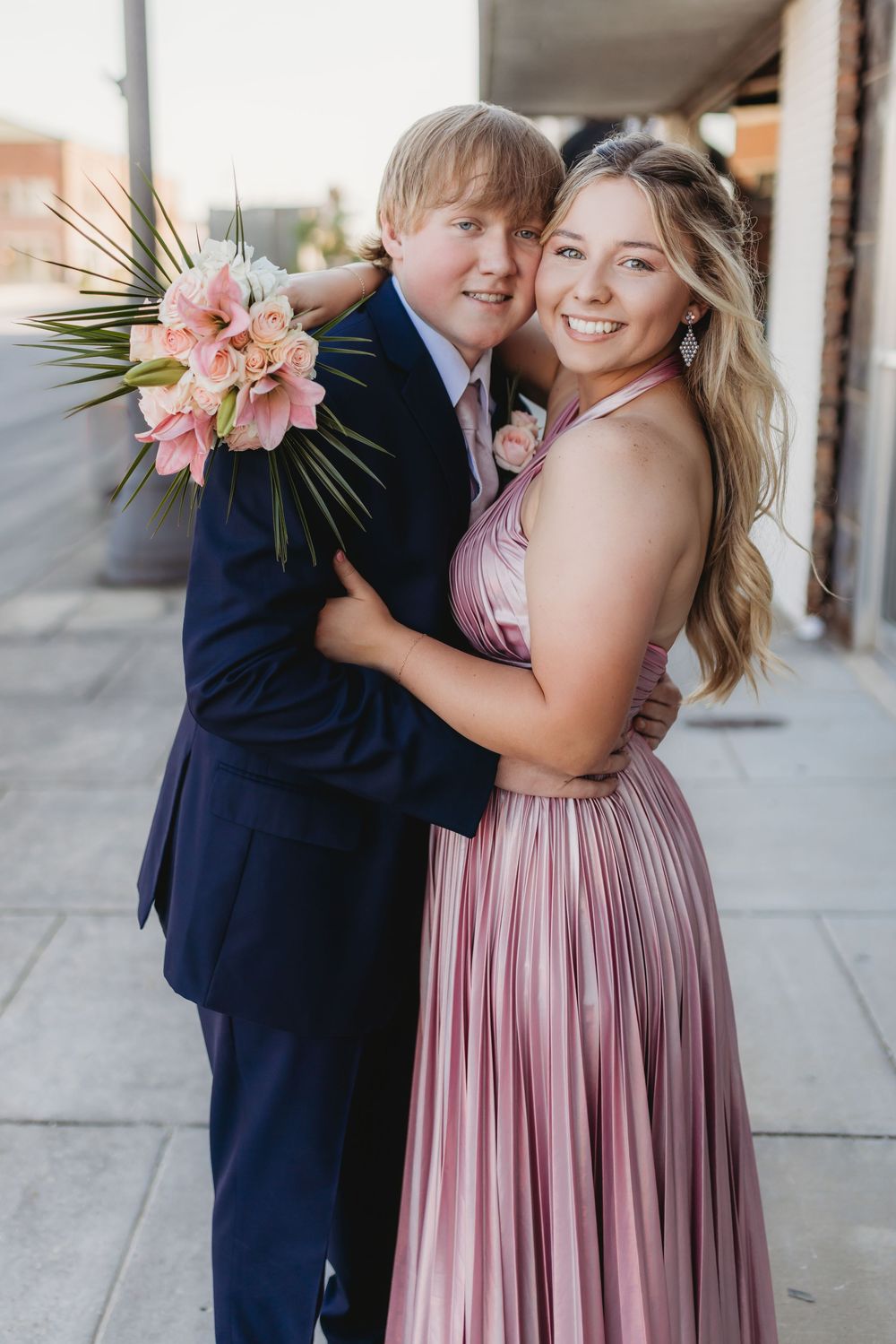 Smiling prom couple embraces on sidewalk, girl in pink pleated dress holds floral bouquet.