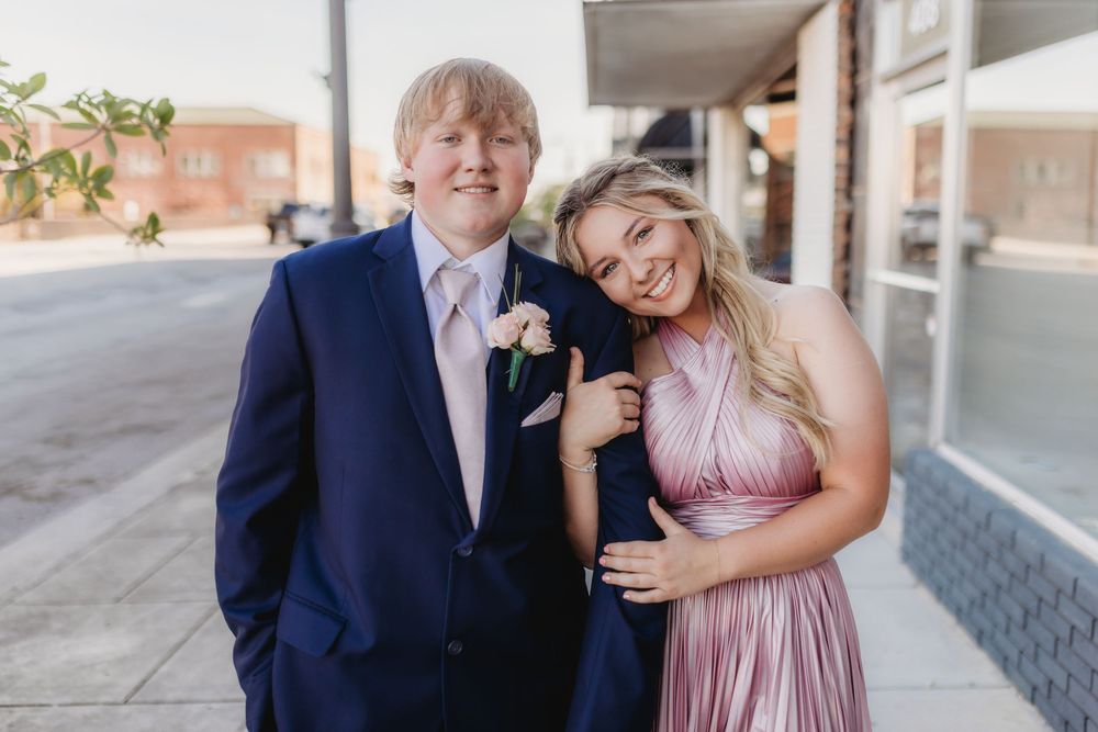 A young couple dressed in formal prom attire, posing outdoors on a sunny day.