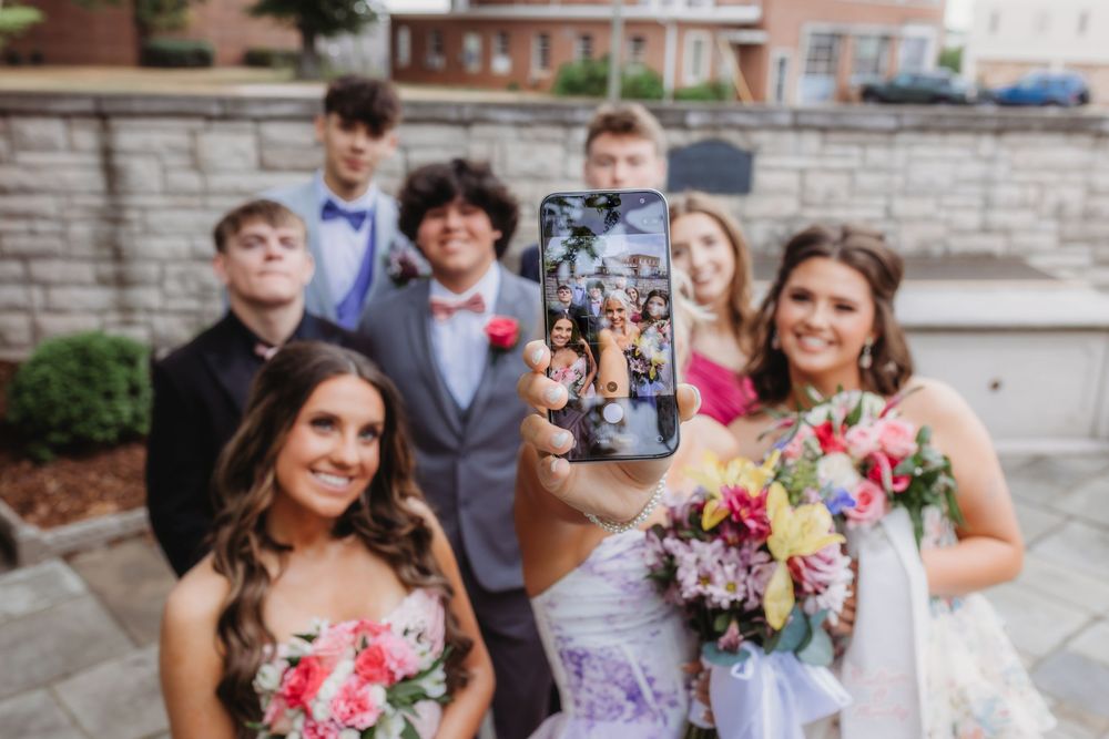 Group of prom-goers posing outdoors while one holds up a phone displaying their photo collage, with girls holding colorful bouquets.