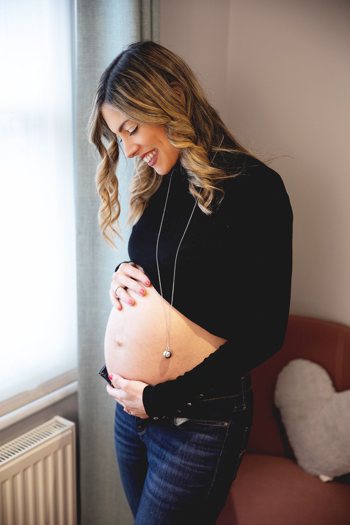 Smiling pregnant woman in black top and jeans cradling her bare baby bump while standing by a window.