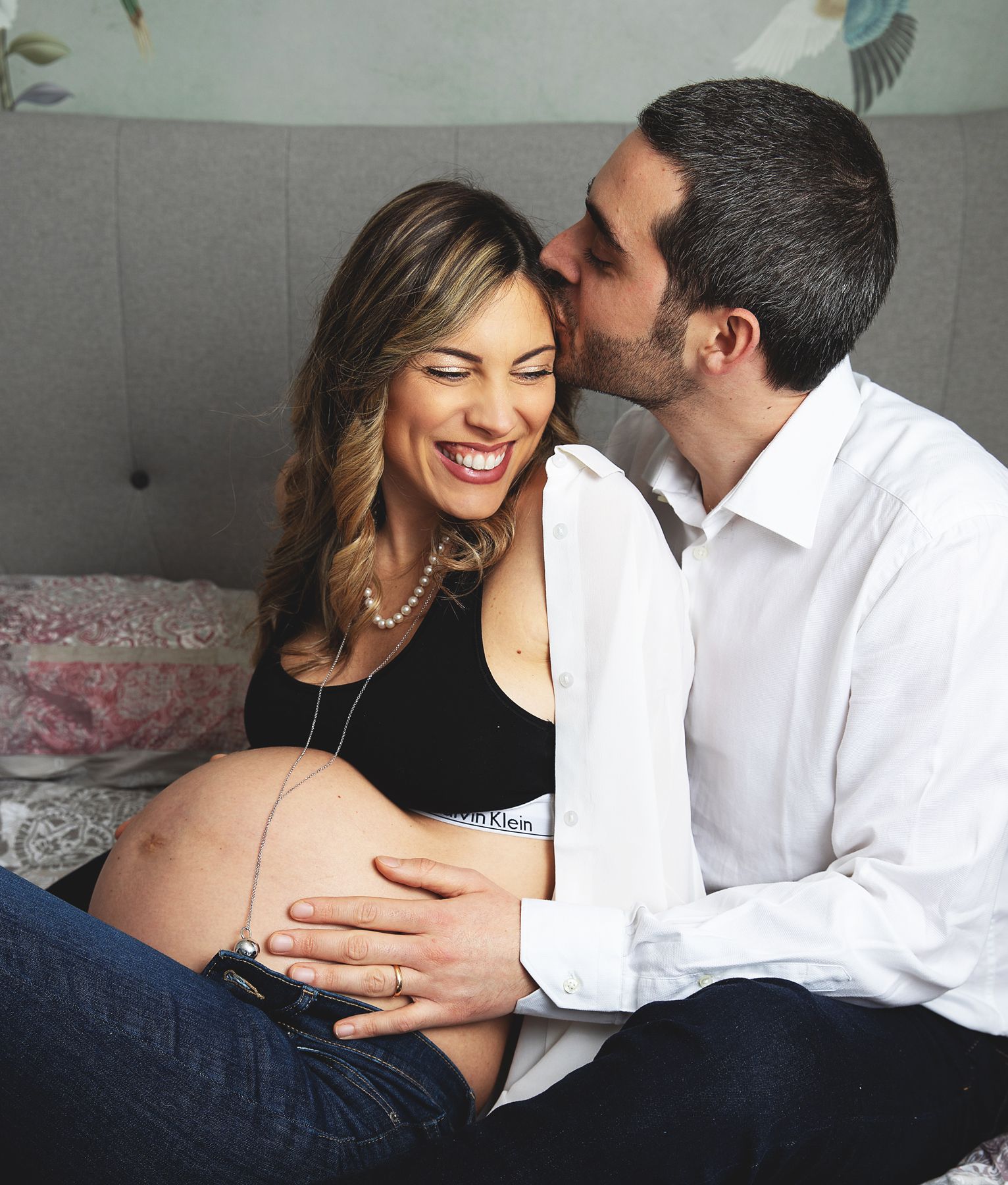 Happy pregnant couple sitting on bed, man kissing woman's cheek while they both cradle her baby bump.