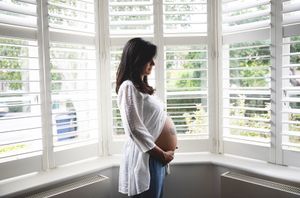 Pregnant woman in white shirt cradling baby bump while standing by bay window with shutters.