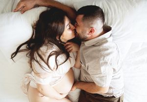 Pregnant couple lying on white bed sharing a tender kiss, captured from above in soft natural light.