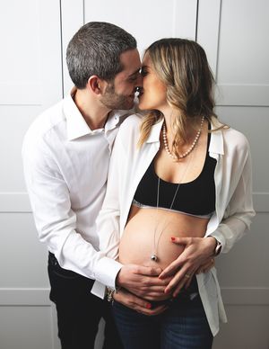 Couple sharing a tender kiss during a maternity photoshoot, man embracing pregnant woman's bare belly indoors.