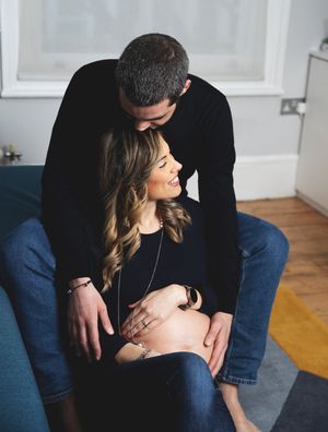 Couple sharing tender moment, man in black sweater embracing smiling pregnant woman in jeans on couch.
