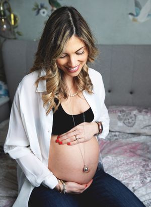 Smiling pregnant woman in white blazer and black crop top cradling baby bump while sitting on bed.