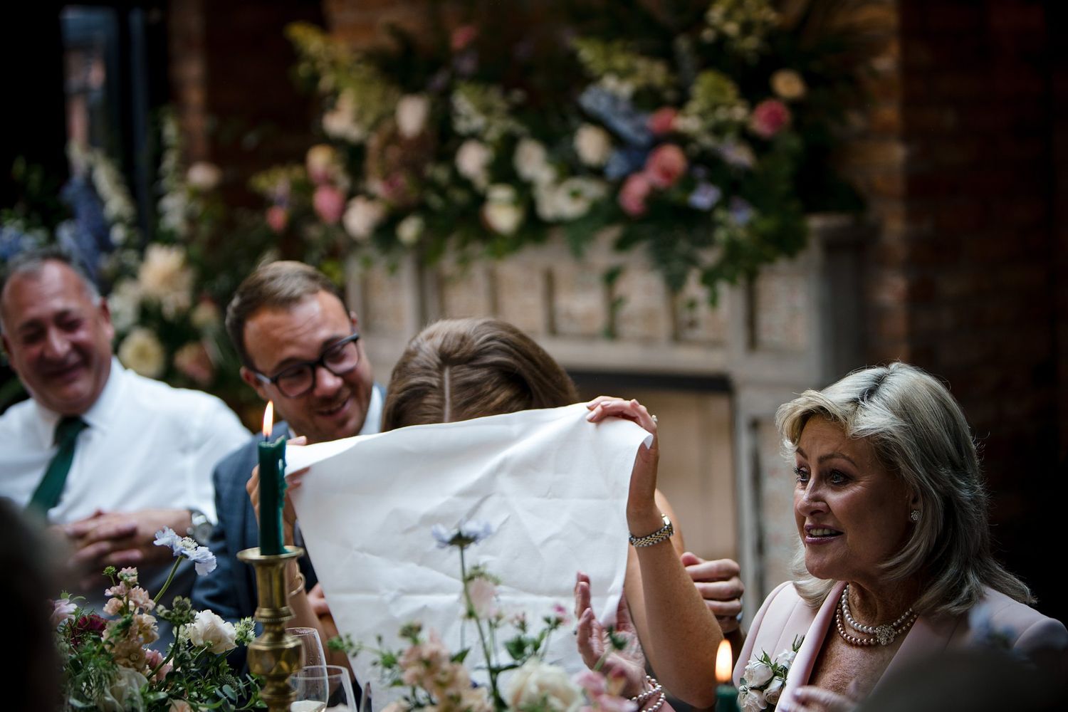 Guests at a wedding reception examine a white cloth near floral centerpieces and candlelight.