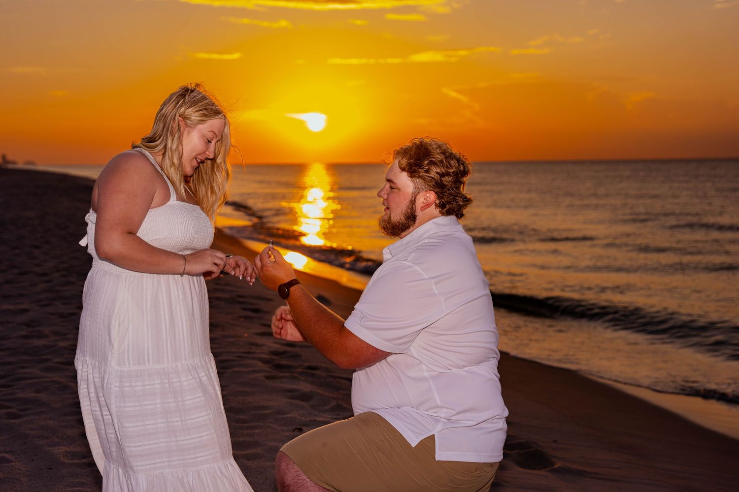 Man proposes to woman on beach at sunset, both in white, with golden sky and ocean in background.