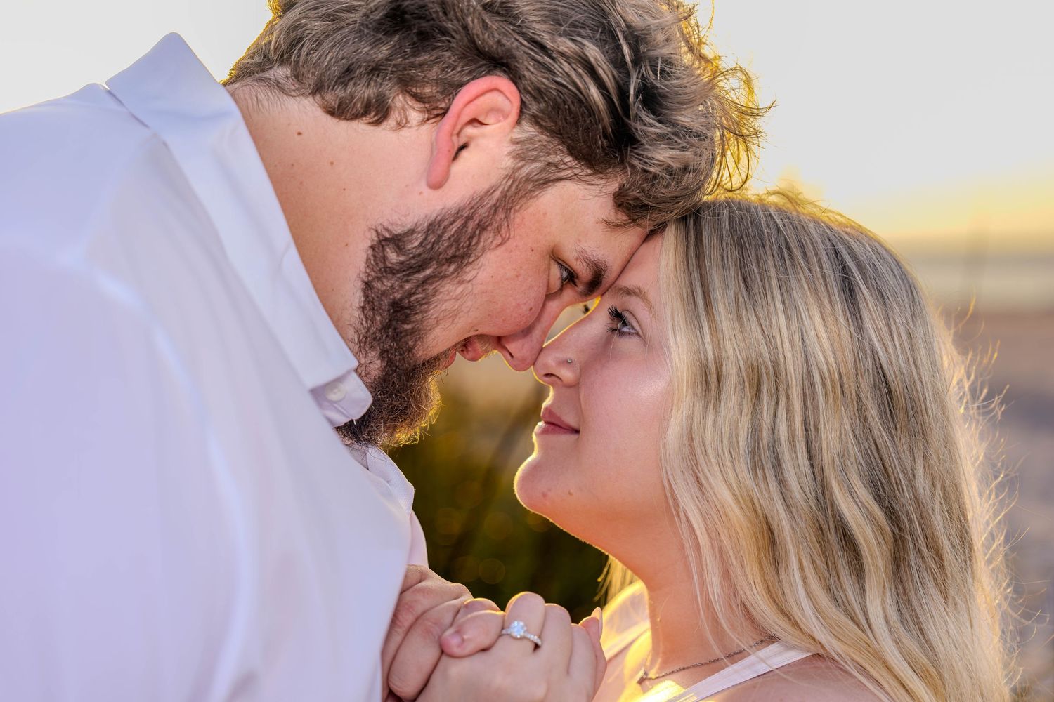 Couple sharing a romantic forehead-to-forehead moment at sunset, woman displaying engagement ring.