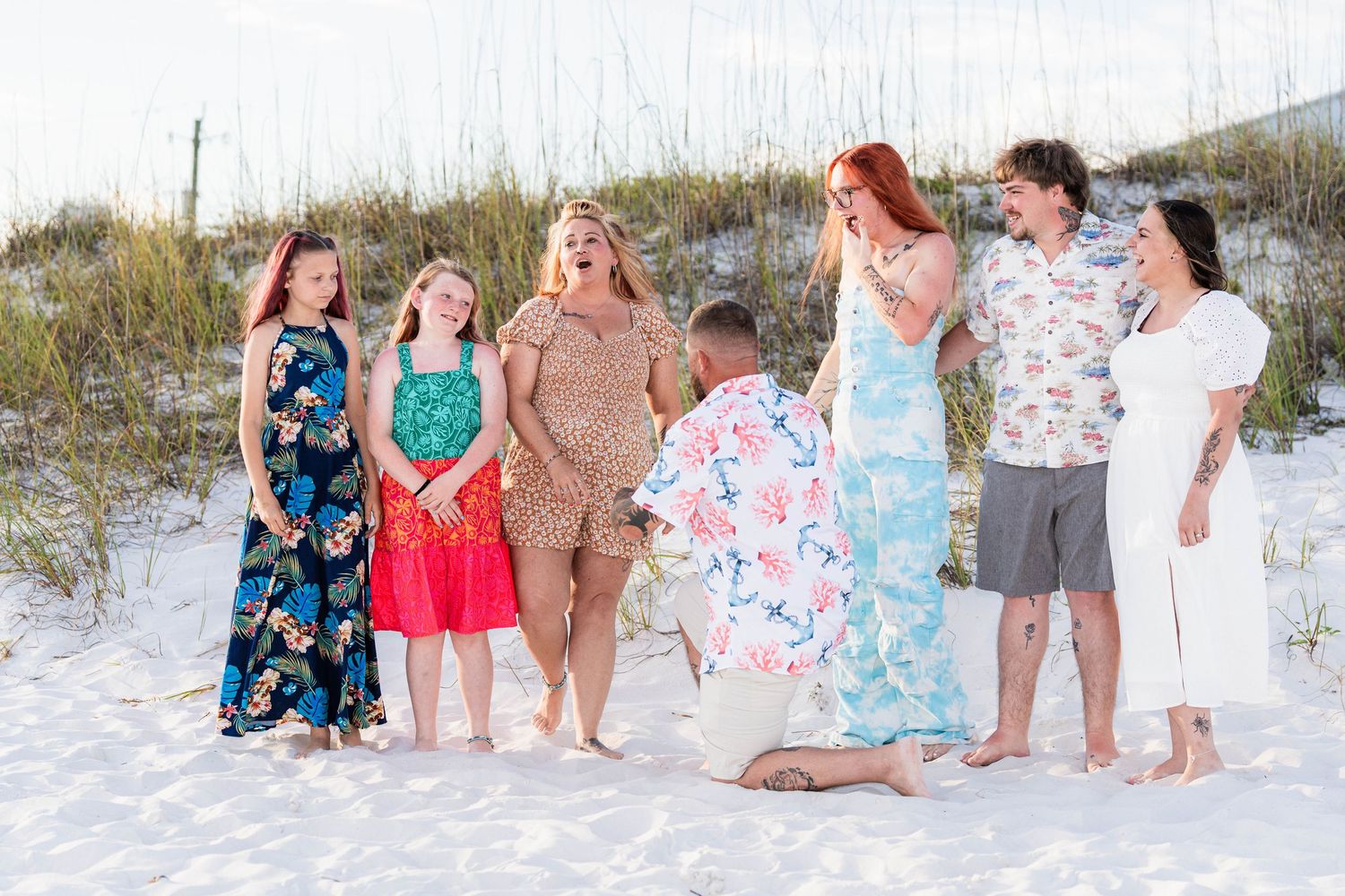 A man proposes on a white sand beach surrounded by family, with sand dunes in the background.