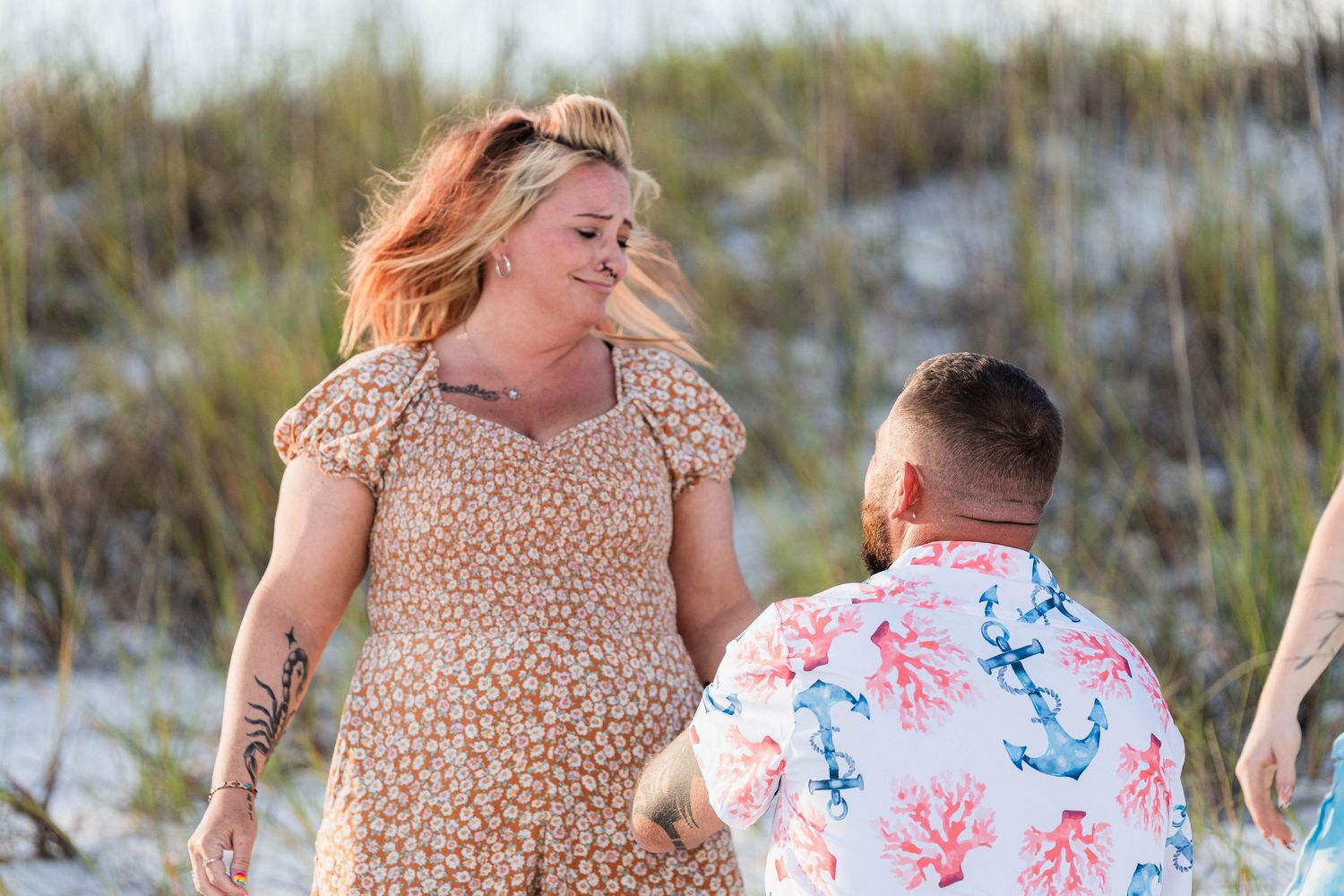Couple holding hands on sandy beach, woman in floral dress with tattoos, man in coral print shirt, dunes in background.